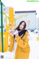 A woman in a yellow coat and hat standing in the snow.